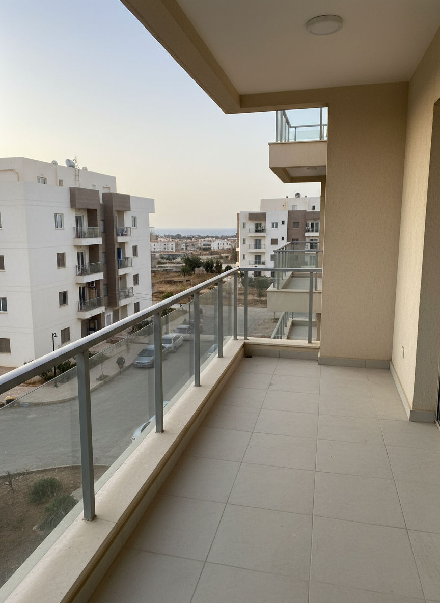 Vue photographique réaliste d’un balcon d’appartement en location à Mostaganem, avec garde-corps en aluminium gris et verre, sol en carrelage beige clair antidérapant. Le balcon surplombe un quartier résidentiel calme avec quelques immeubles modernes et un aperçu de la mer à l’horizon, légèrement floutés pour garder le focus sur l’espace extérieur. Lumière de matinée, douce, faisant briller discrètement les surfaces métalliques et soulignant la propreté des lieux. Prise de vue à niveau du regard, cadrage horizontal montrant la longueur du balcon et une partie de la façade. Ambiance tranquille et agréable, idéale pour mettre en avant l’atout extérieur d’un bien immobilier.